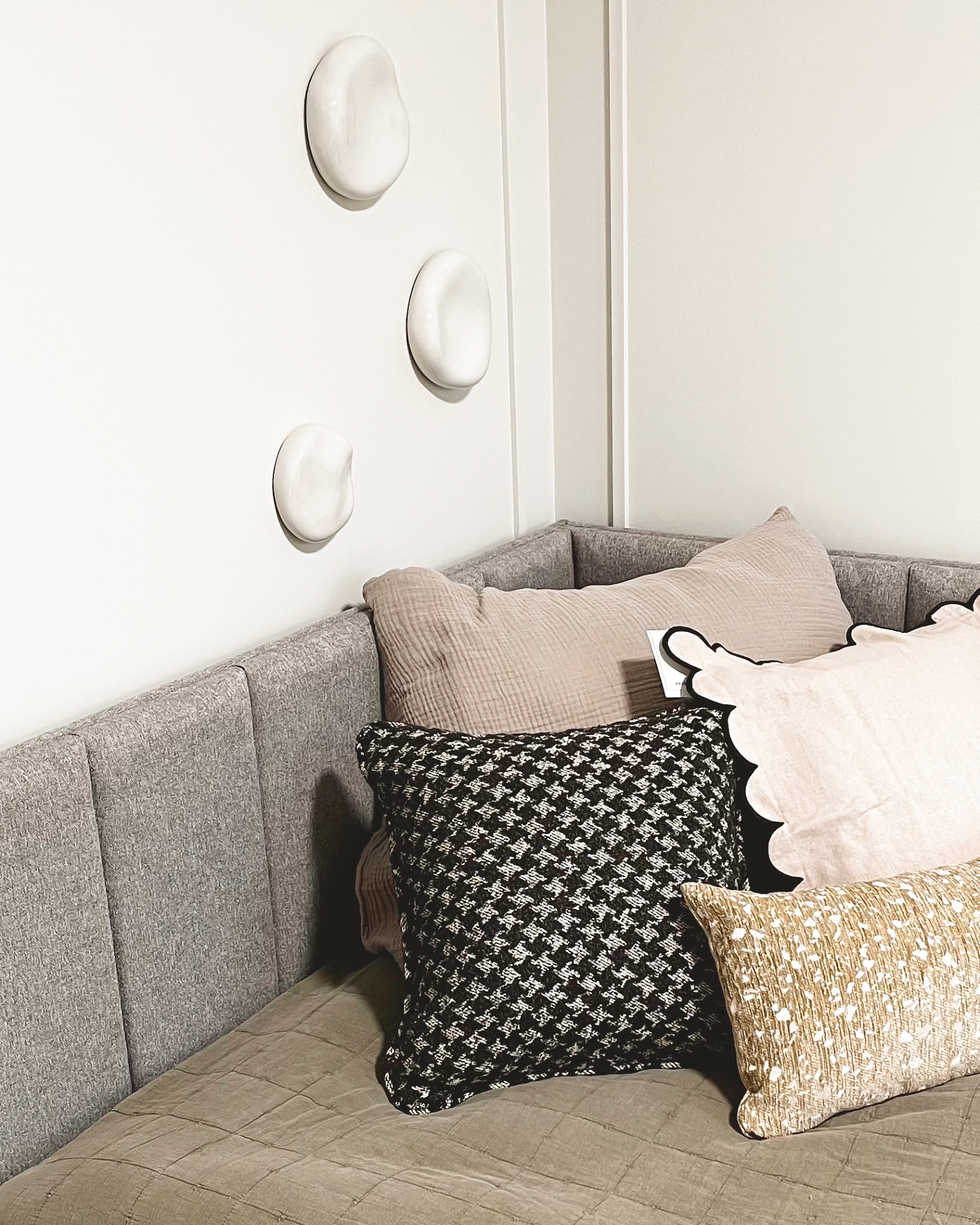Stylowy narożnik z szarą tapicerką, zdobiony poduszkami o różnych wzorach i fakturach. Na ścianie dekoracyjne, białe, ceramiczne elementy.