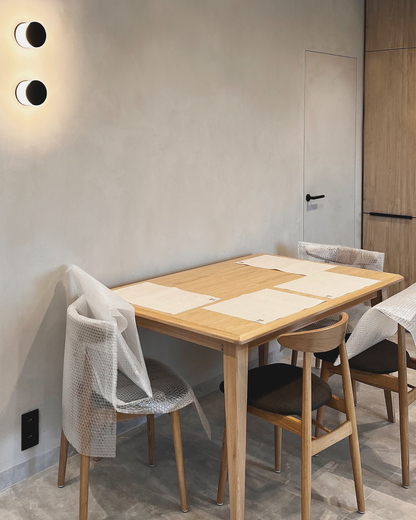 Jasne, minimalistyczne wnętrze: drewniany stół z krzesłami częściowo owiniętymi folią ochronną. Na stole podkładki. W tle ściana z oświetleniem i ukryte drzwi.