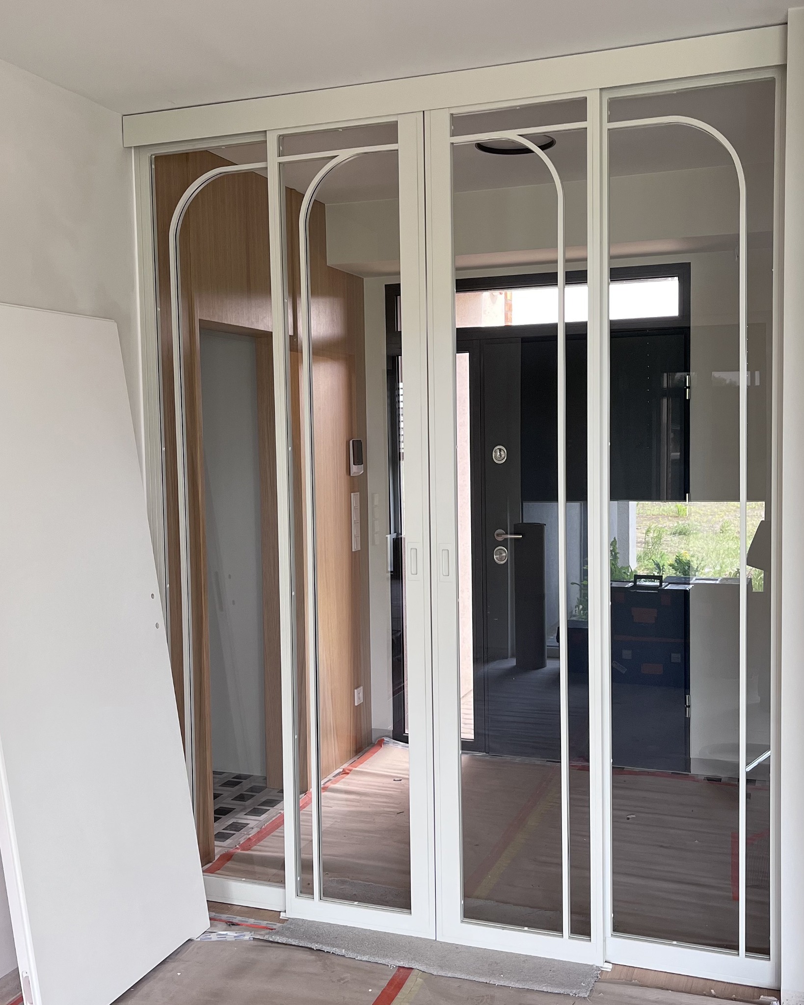Białe, przesuwne drzwi z łukowatymi elementami w szklanych panelach, widoczne w trakcie montażu. W tle widoczne inne pomieszczenia i drzwi wejściowe.