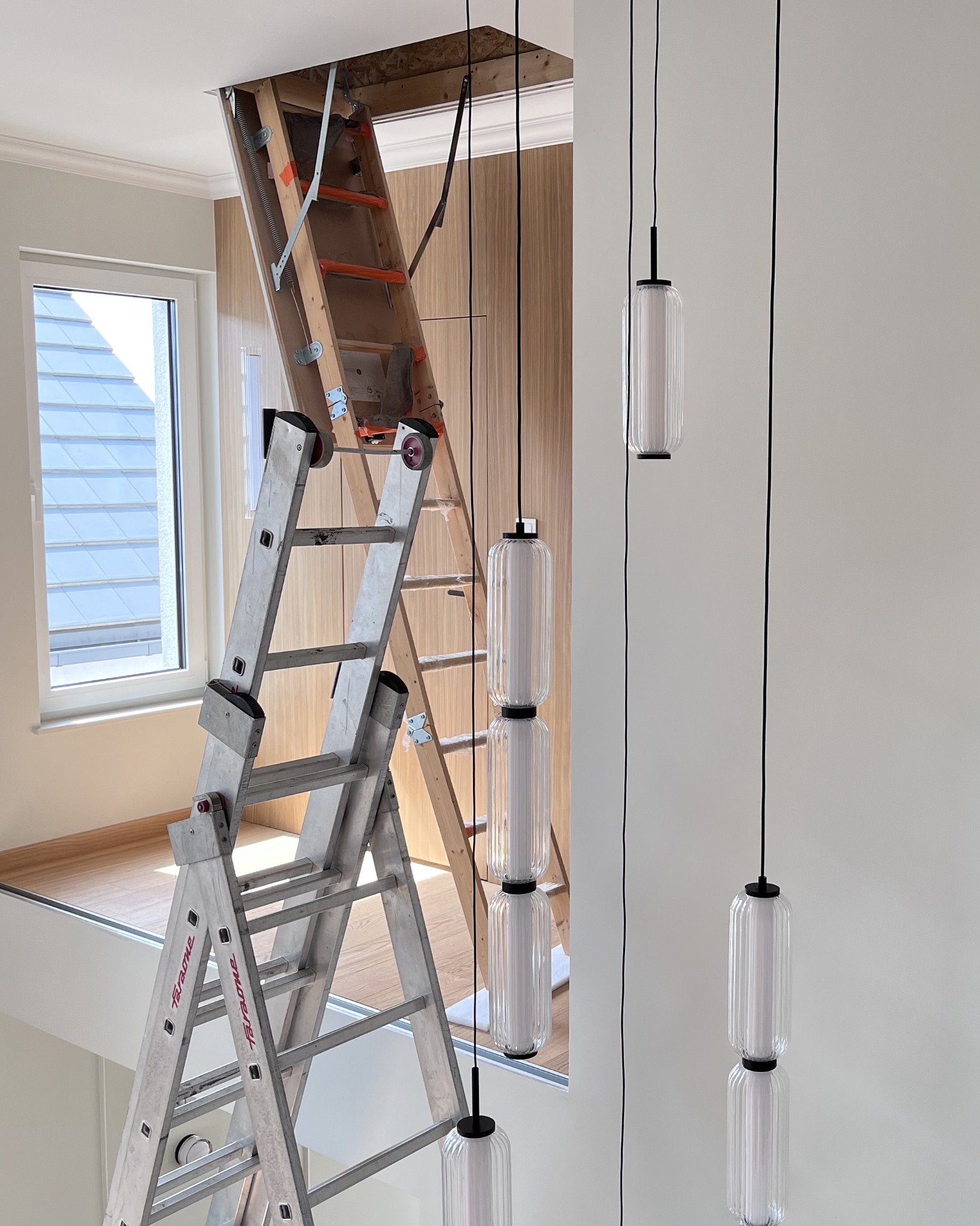 Montaż lamp wiszących w nowym wnętrzu. Aluminiowa drabina oparta o schody strychowe. Widoczne elementy wykończenia i okno. Neutralna kolorystyka ścian.