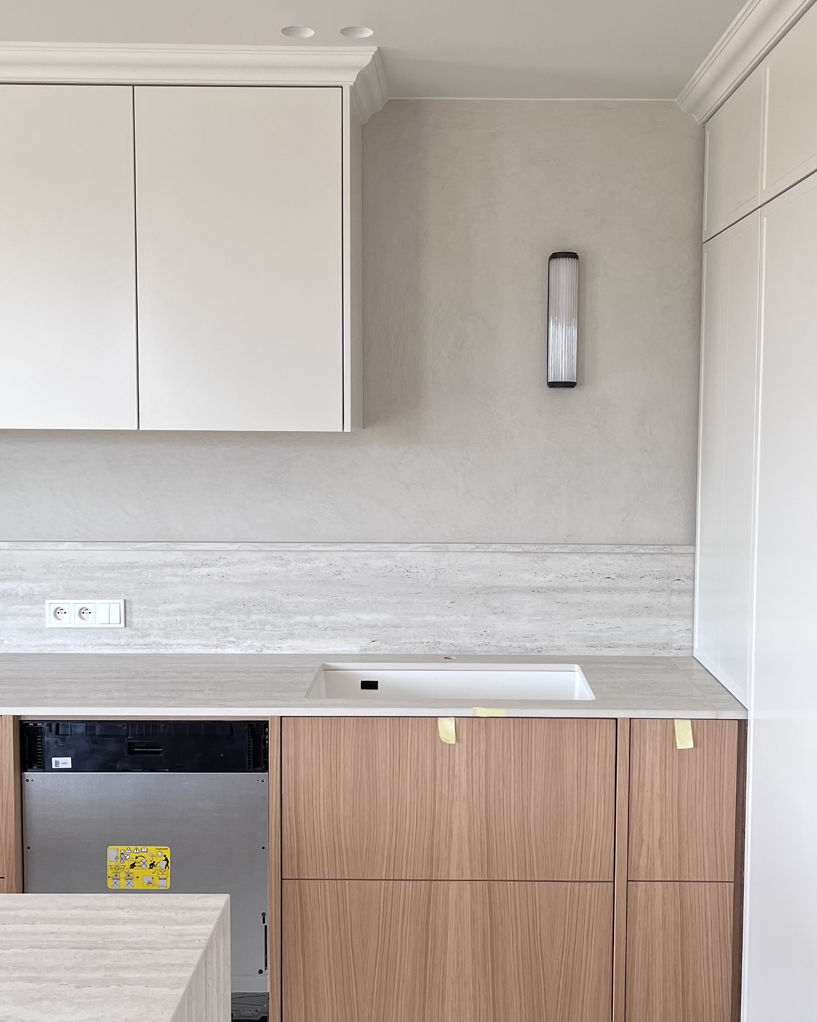 Jasna kuchnia po remoncie: białe szafki górne z dekoracyjną listwą, drewniane szafki dolne, blat z szarego kamienia, podłużna lampa ścienna. Widoczny fragment zmywarki.