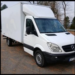 Zapała Sławomir - Biały samochód dostawczy Mercedes-Benz Sprinter z zabudową kontenerową, zaparkowany na asfalcie. Widoczne drzewa w tle. Tablica rejestracyjna KNT-09.
