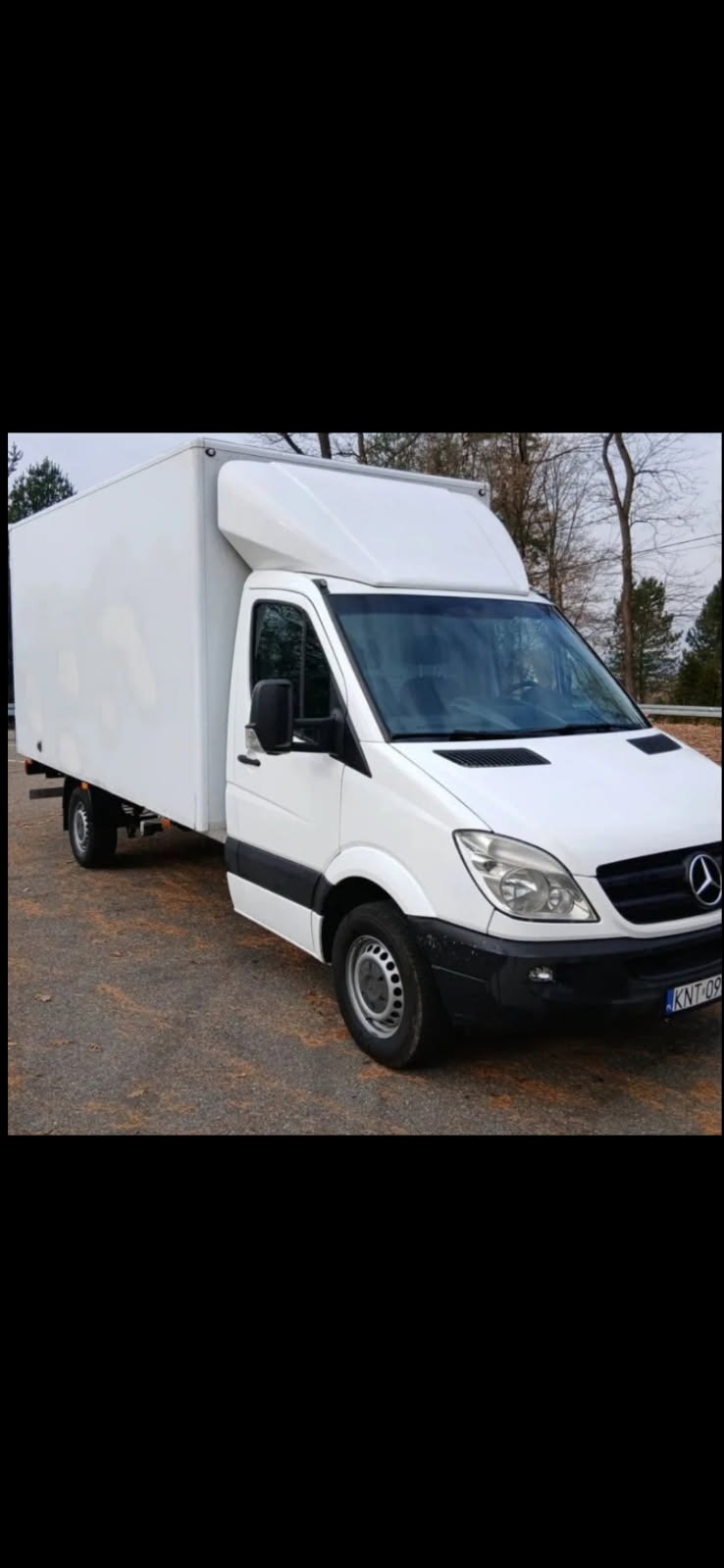 Biały samochód dostawczy Mercedes-Benz Sprinter z zabudową kontenerową, zaparkowany na asfalcie. Widoczne drzewa w tle. Tablica rejestracyjna KNT-09.