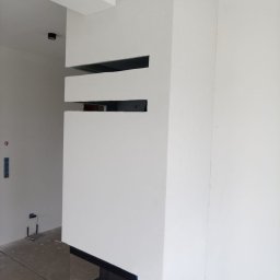 STEPAN HOI - Biała ściana z wnękami i kominkiem w minimalistycznym stylu. Widoczne niedoskonałości wykończenia, surowy beton na podłodze. Ujęcie z dołu, podkreślające pionowość.