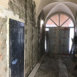 STEPAN HOI - Korytarz w trakcie prac wykończeniowych: ściany w surowym tynku, zabezpieczone drzwi folią, widoczne instalacje elektryczne, łukowe przejście z oknem na końcu.
