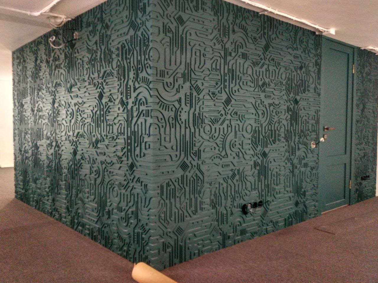 Pokój z unikalną, teksturowaną ścianą w odcieniach zieleni, przypominającą płytę główną komputera. Widoczne gniazdka elektryczne i dopasowane kolorystycznie drzwi.