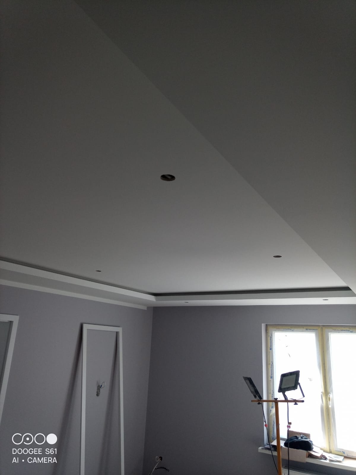 Szare wnętrze po malowaniu ścian i sufitu z ozdobną sztukaterią. Widoczne lampy robocze i okno z widokiem na zewnątrz. Minimalistyczny design.