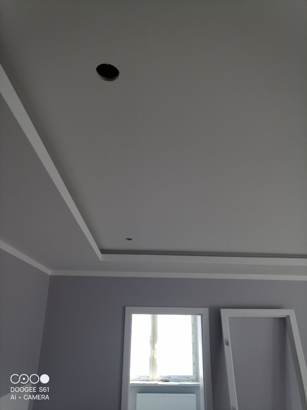 Szare ściany i sufit z dekoracyjnymi białymi listwami. Widoczne otwory na oświetlenie i okno z białymi ramami. Częściowo widoczna ościeżnica drzwi.