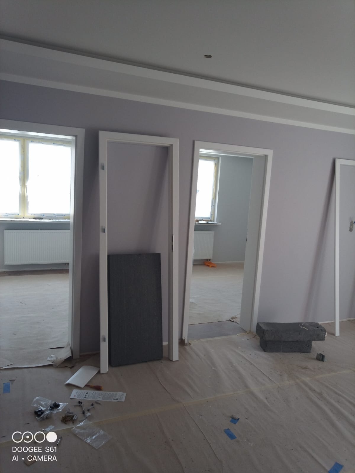 Wnętrze w trakcie remontu: trzy białe ościeżnice drzwiowe, podłoga zabezpieczona folią, widoczne akcesoria montażowe. Ściany pomalowane na jasny fiolet.