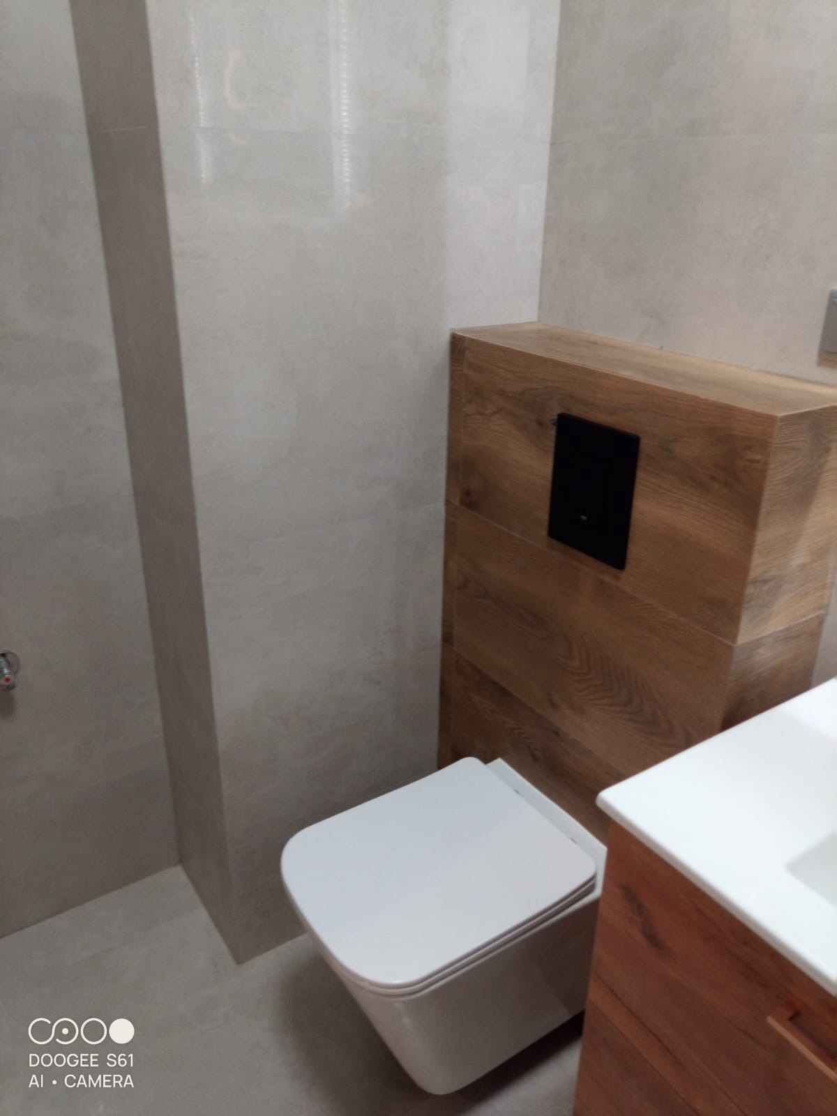 Nowoczesna toaleta podwieszana z drewnianą obudową spłuczki i szarą glazurą w wykończonej łazience. Minimalistyczny design, widok z boku.