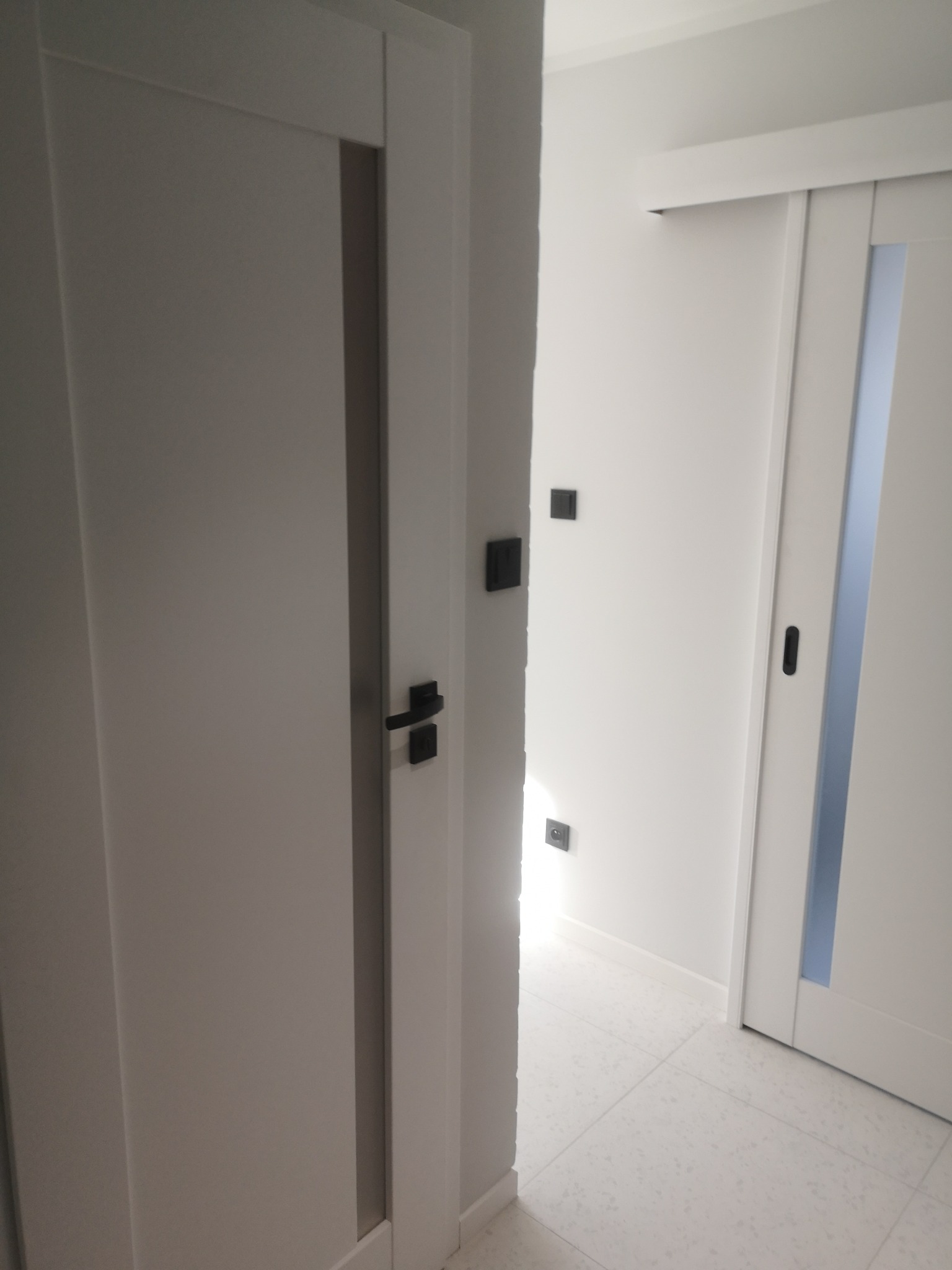 Nowoczesne, białe drzwi z czarną klamką w minimalistycznym wnętrzu. Widoczne gniazdka i włączniki. W tle przesuwne drzwi z szybą.