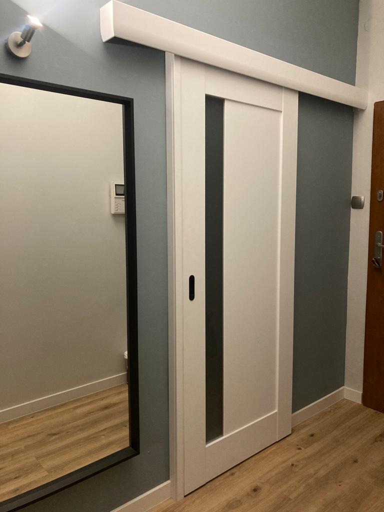 Białe, przesuwne drzwi z czarnymi, pionowymi wstawkami, zamontowane w szarej ścianie. Obok widoczne lustro w czarnej ramie i fragment drewnianej podłogi.