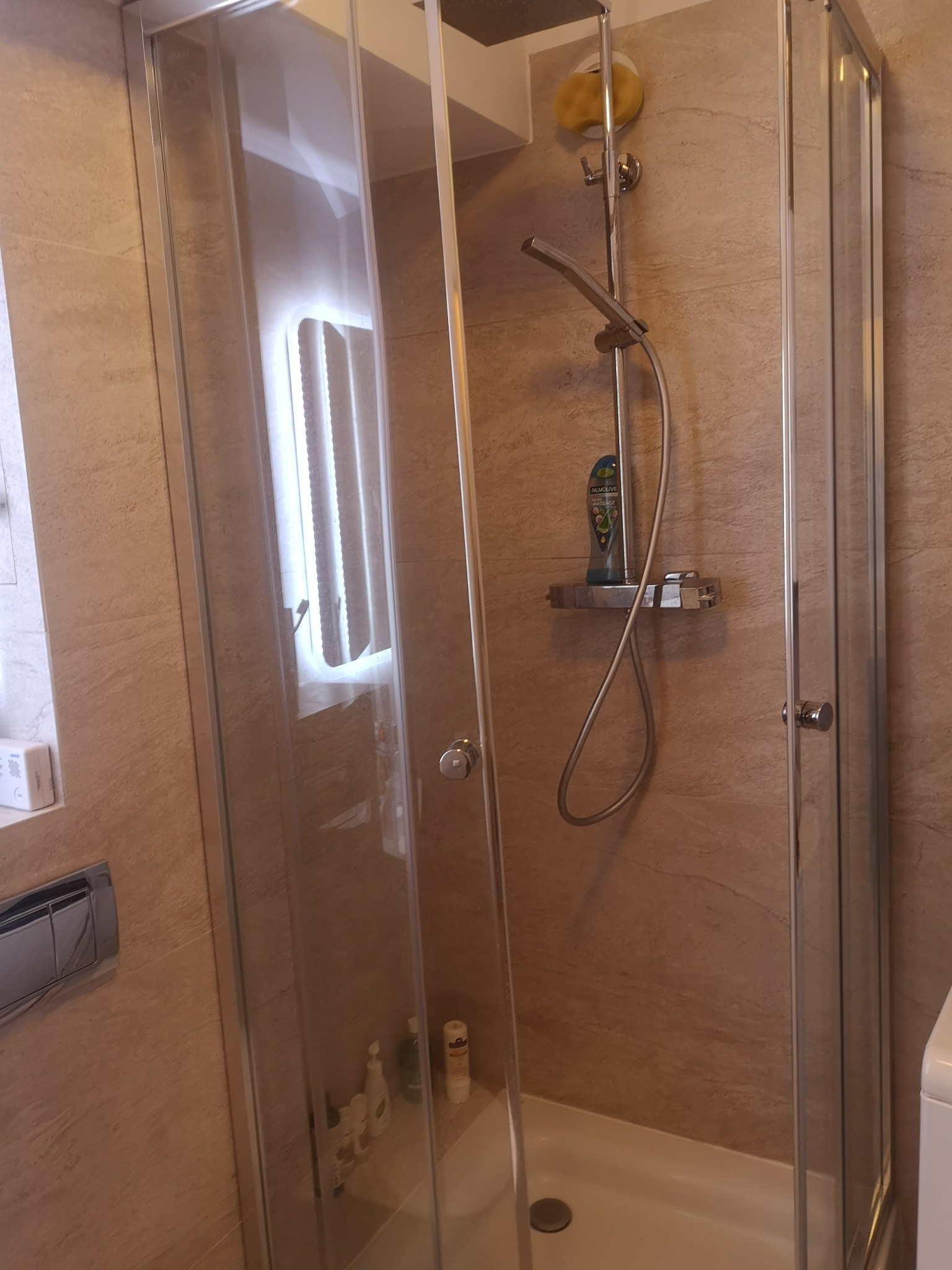 Kabina prysznicowa z chromowanymi profilami i beżowymi płytkami. Widoczna słuchawka prysznicowa, półka z kosmetykami i detale wykończenia. Ujęcie z wnętrza łazienki.