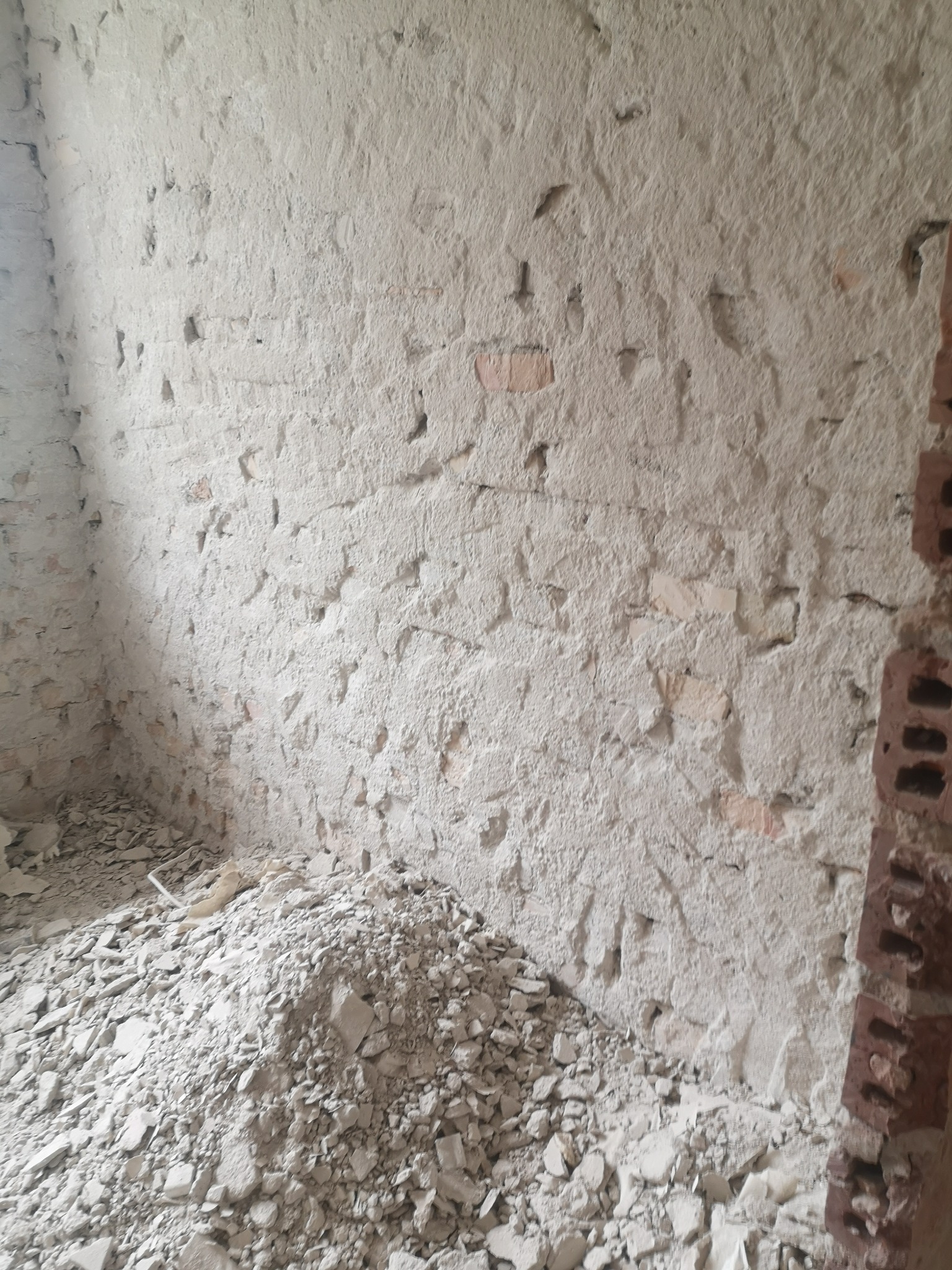 Surowa ściana z cegły i resztki gruzu po skuciu tynku. Widoczne nierówności i uszkodzenia. Fragment ściany z czerwonej cegły po prawej stronie kadru.