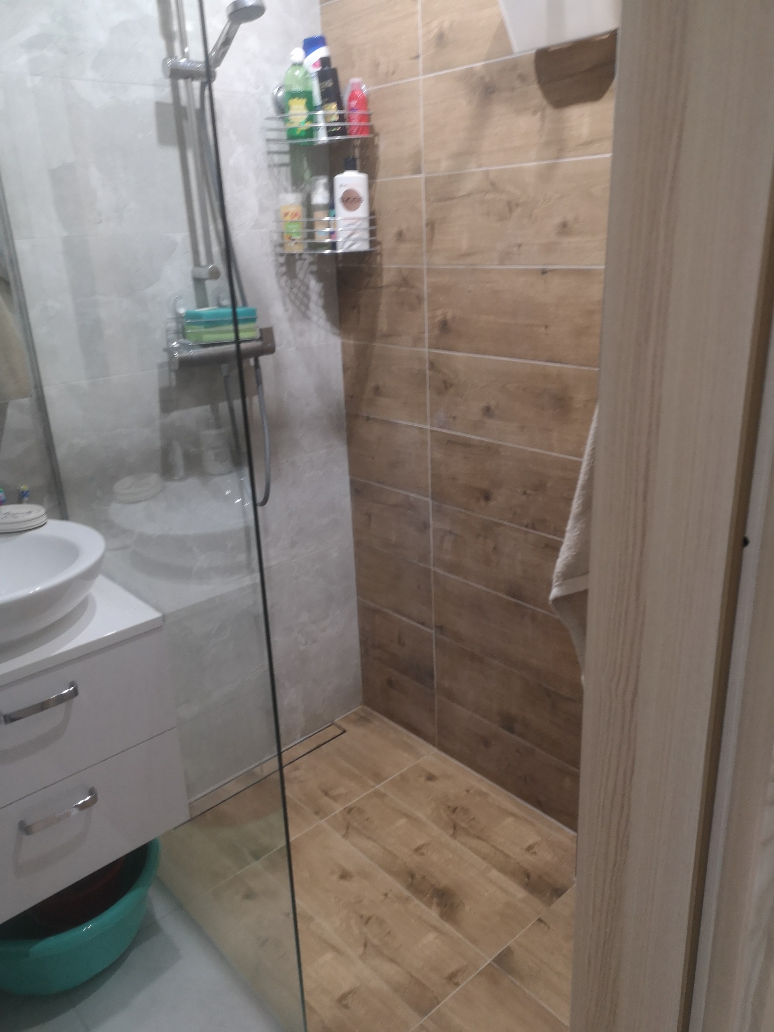 Nowoczesna łazienka z kabiną prysznicową typu walk-in, wykończona płytkami imitującymi drewno. Widoczna umywalka z szafką i chromowana armatura.