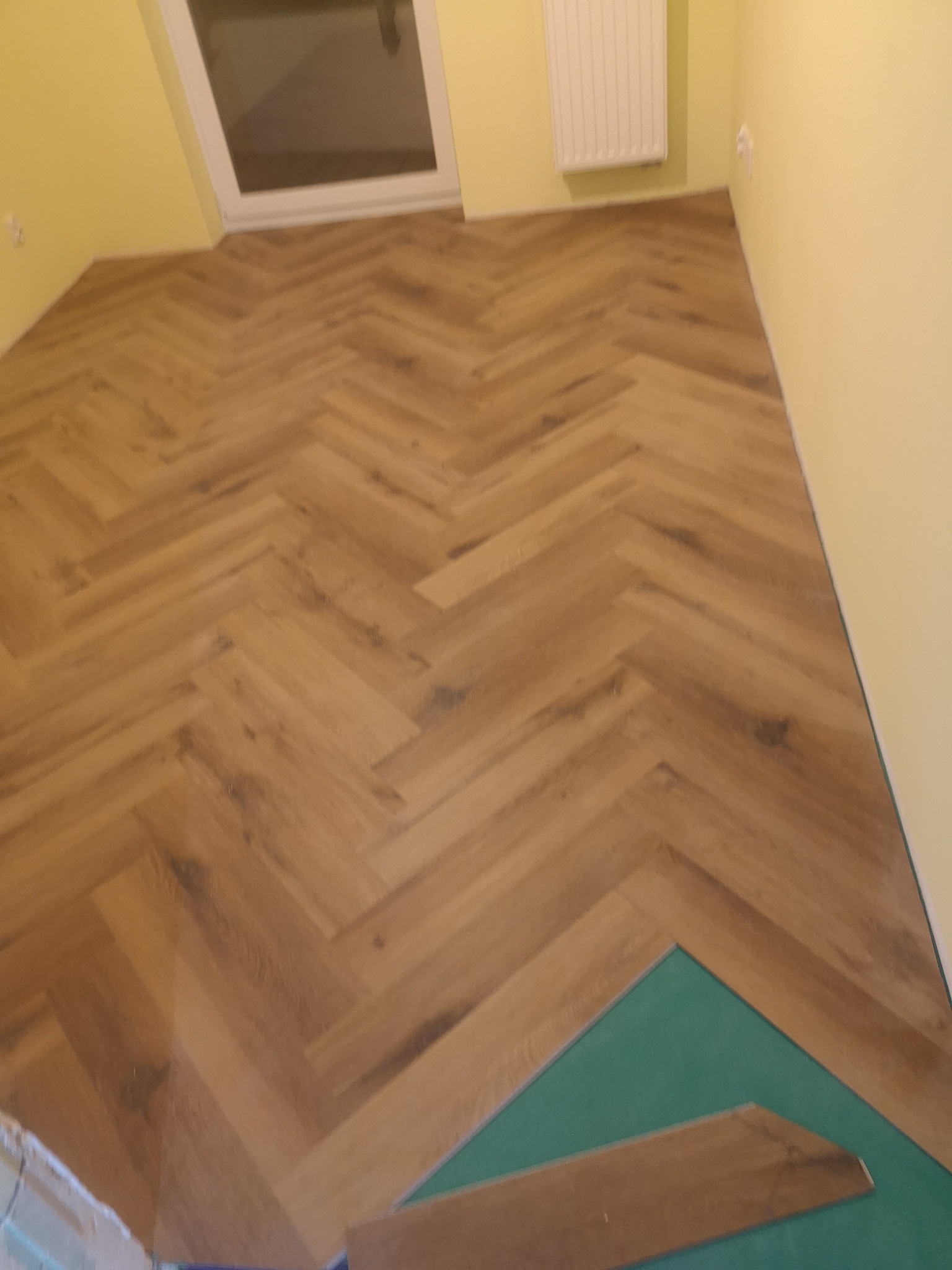 Układanie parkietu w jodełkę w Świnoujściu. Widoczny fragment podkładu i niedokończony fragment podłogi. Żółte ściany i okno dachowe.