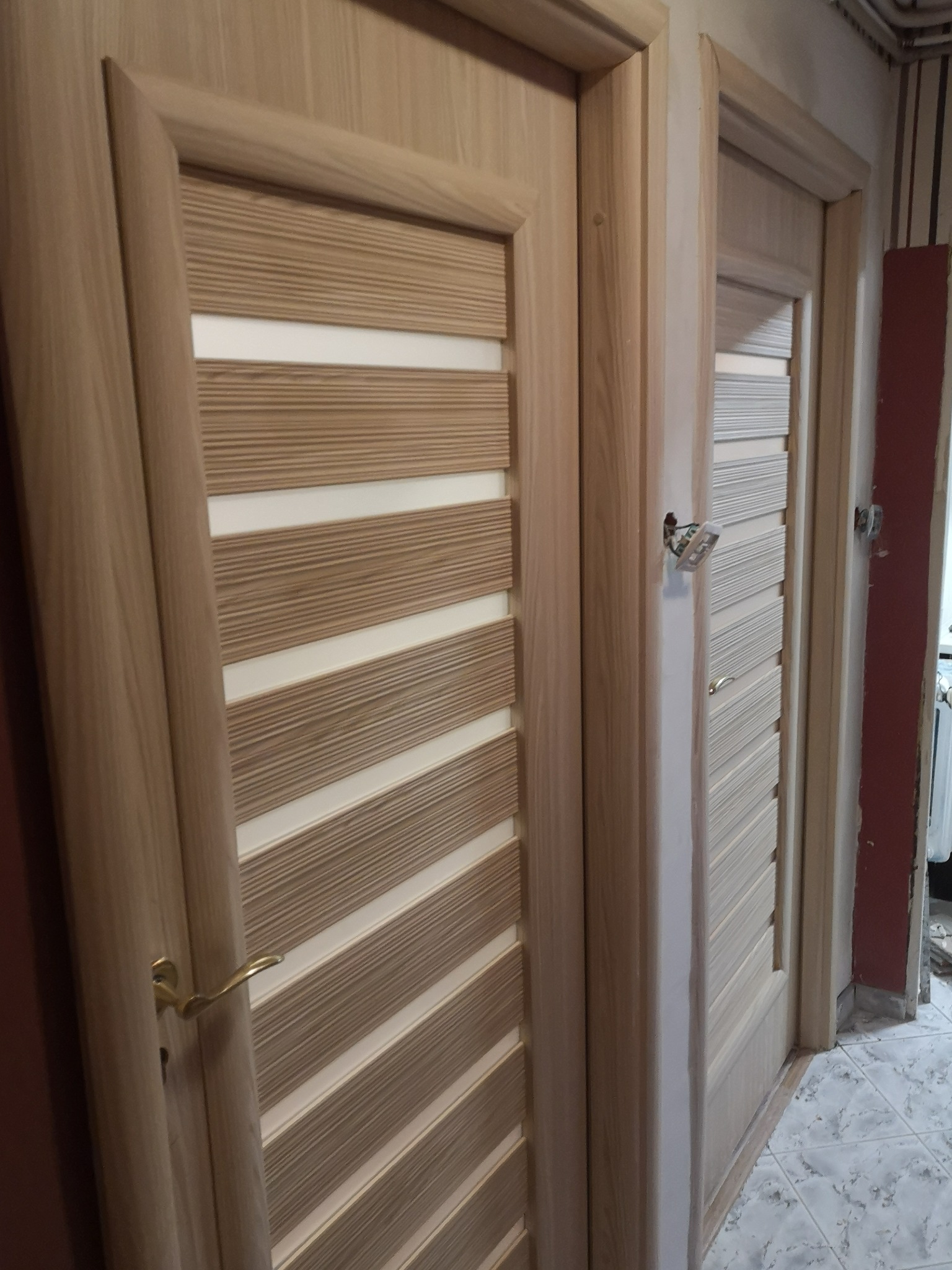 Dwa jasne, drewniane drzwi wewnętrzne z poziomymi panelami, widoczne z bliska. Jeden z zamontowaną klamką, drugi z widoczną puszką elektryczną obok ościeżnicy.