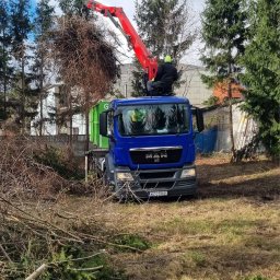 DACAR SPÓŁKA Z OGRANICZONĄ ODPOWIEDZIALNOŚCIĄ - Niebieska ciężarówka MAN z dźwigiem i pracownikiem podczas wycinki drzew. W tle zielony kontener i drzewa iglaste. Na ziemi leżą ścięte gałęzie.
