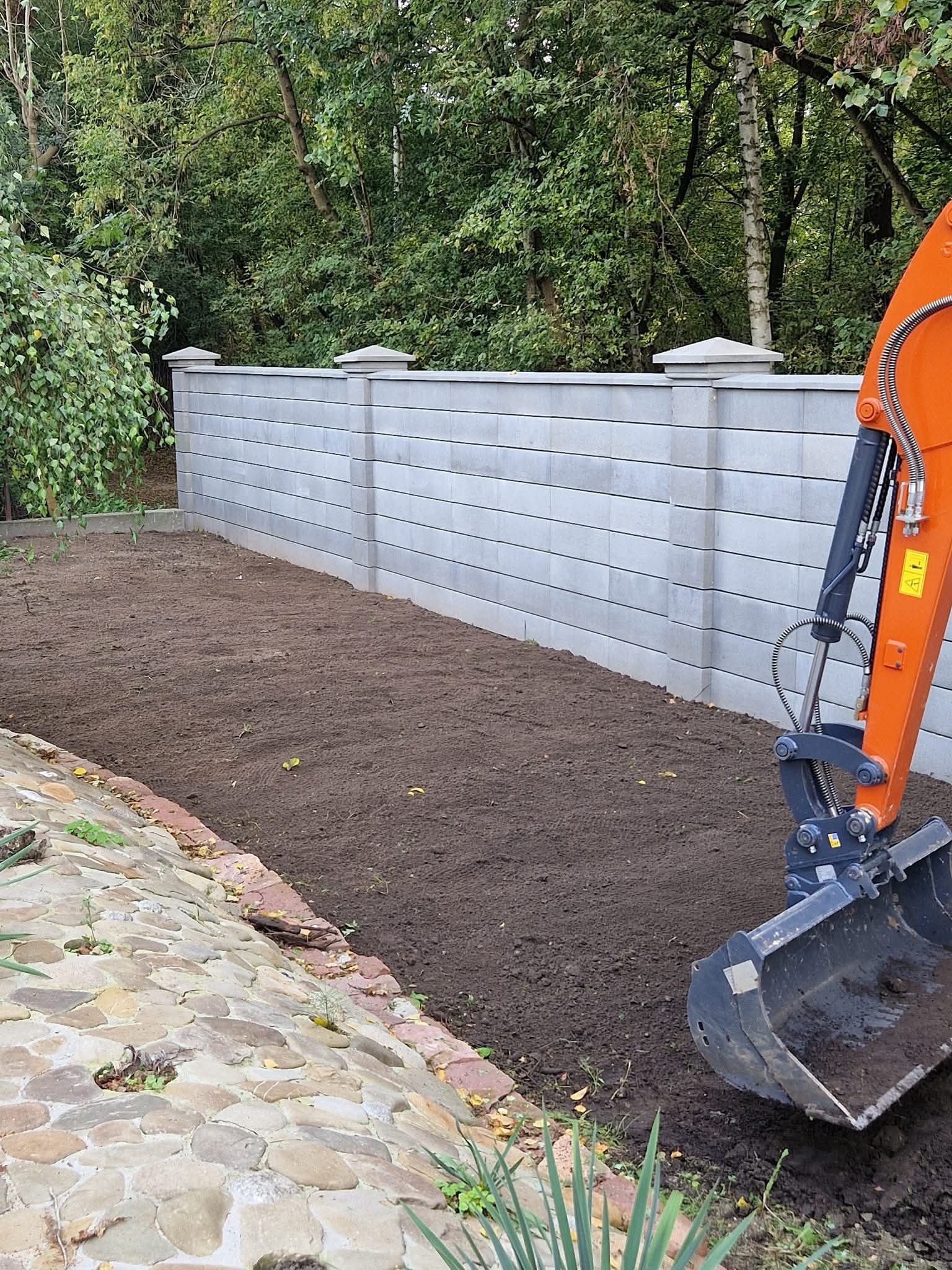 Nowoczesny, szary mur oporowy z betonowych bloczków, świeżo wyrównana ziemia i ramię koparki. Tło stanowią drzewa.