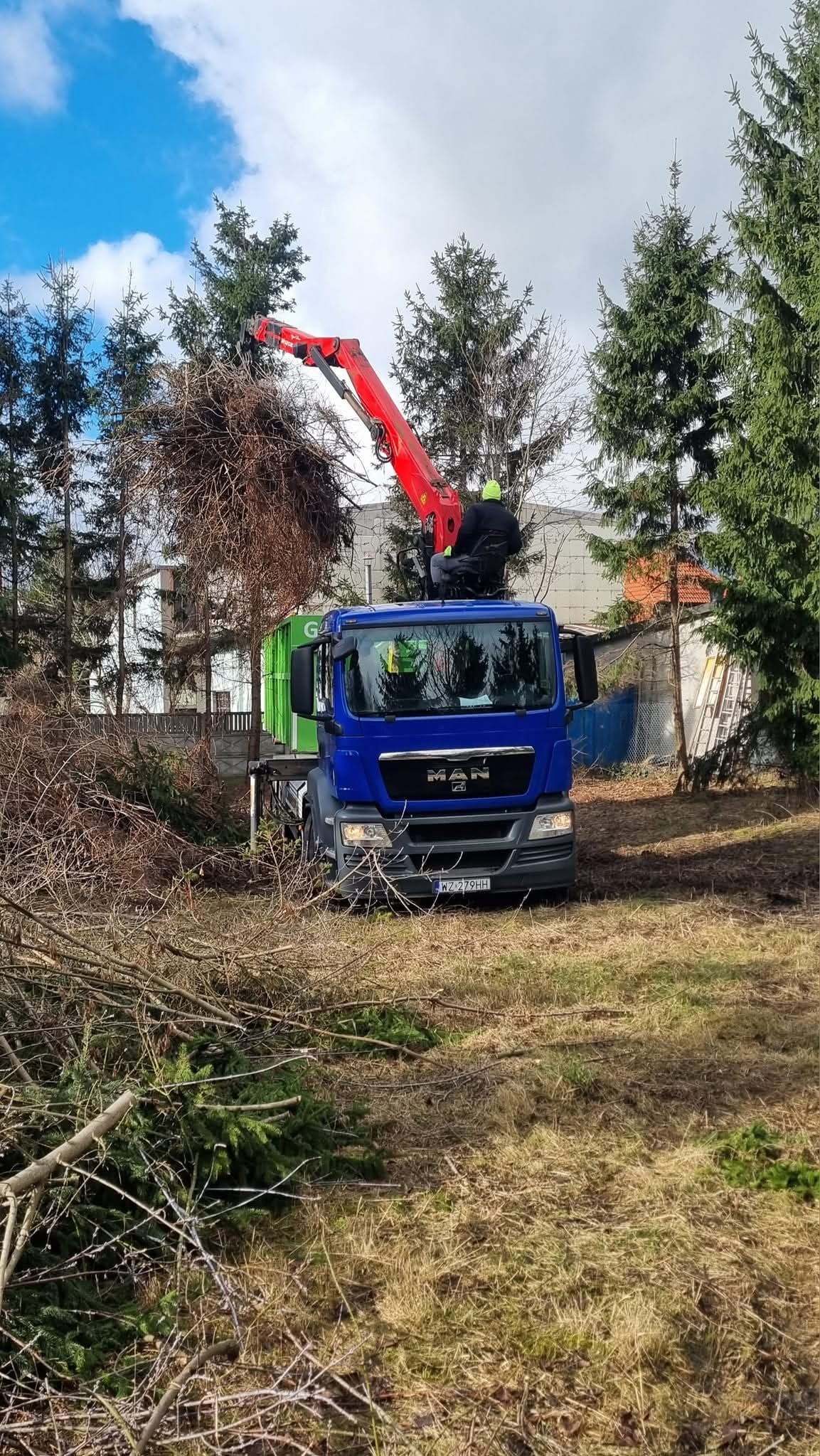 Niebieska ciężarówka MAN z dźwigiem i pracownikiem podczas wycinki drzew. W tle zielony kontener i drzewa iglaste. Na ziemi leżą ścięte gałęzie.