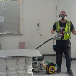 Wykończenia Fot-mar S.A. - Pracownik w masce i kamizelce trzyma szlifierkę do gipsu w pomieszczeniu z płytami gipsowymi i odkurzaczem. Widoczne kable i niedokończone ściany.