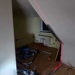 Wykończenia Fot-mar S.A. - Skośny sufit poddasza w Szczecinie, wyznaczanie poziomów laserem. Widoczne narzędzia, płyta gipsowa i odkurzacz przemysłowy. Przygotowanie do montażu.