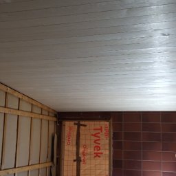 Wykończenia Fot-mar S.A. - Montaż paneli sufitowych z widoczną konstrukcją nośną i izolacją. Fragment ściany z brązowymi płytkami. Pracownik w trakcie montażu. Otwór na oświetlenie w suficie.