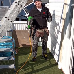 Wykończenia Fot-mar S.A. - Uśmiechnięty monter z pistoletem pneumatycznym na tle białej elewacji domu. Widoczna drabina, narzędzia i przewody. Dzień słoneczny, praca na zewnątrz.