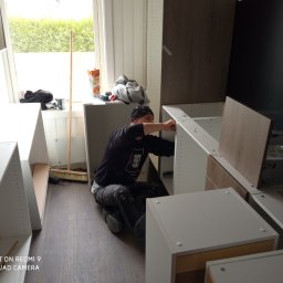 Wykończenia Fot-mar S.A. - Monterażysta w trakcie montażu białych szafek kuchennych w jasnym wnętrzu. Widoczny fragment okna i podłogi. Na blacie leżą narzędzia i napój.
