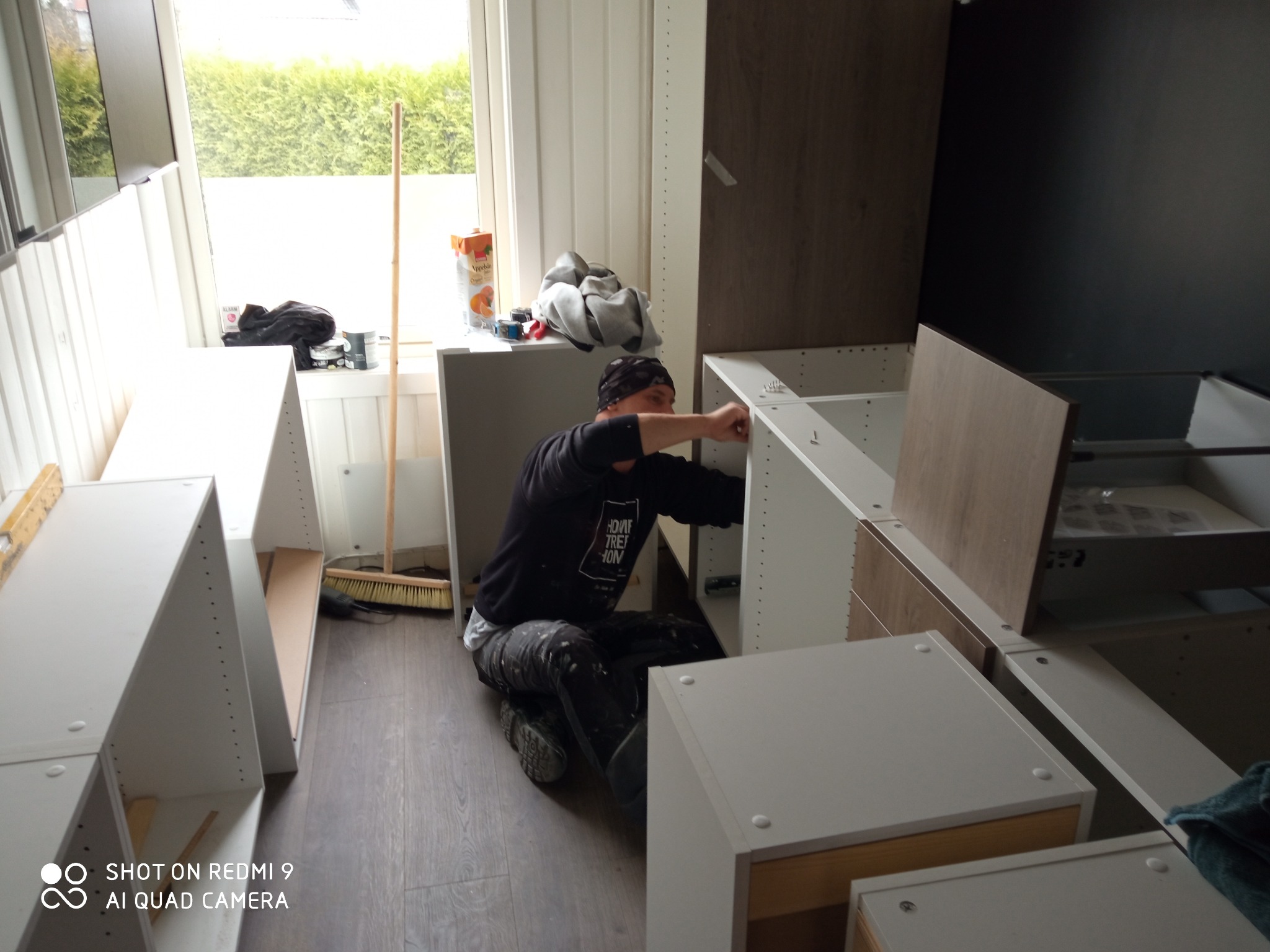 Monterażysta w trakcie montażu białych szafek kuchennych w jasnym wnętrzu. Widoczny fragment okna i podłogi. Na blacie leżą narzędzia i napój.