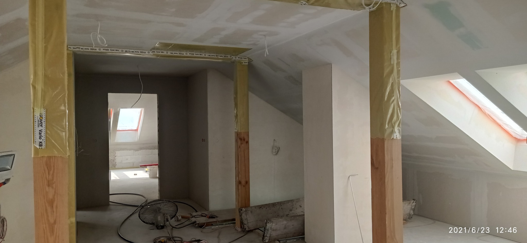 Poddasze w trakcie prac wykończeniowych: widoczne drewniane słupy z folią, płyty karton-gips na suficie, wentylator na podłodze. W tle okno dachowe i surowy stan pomieszczenia.
