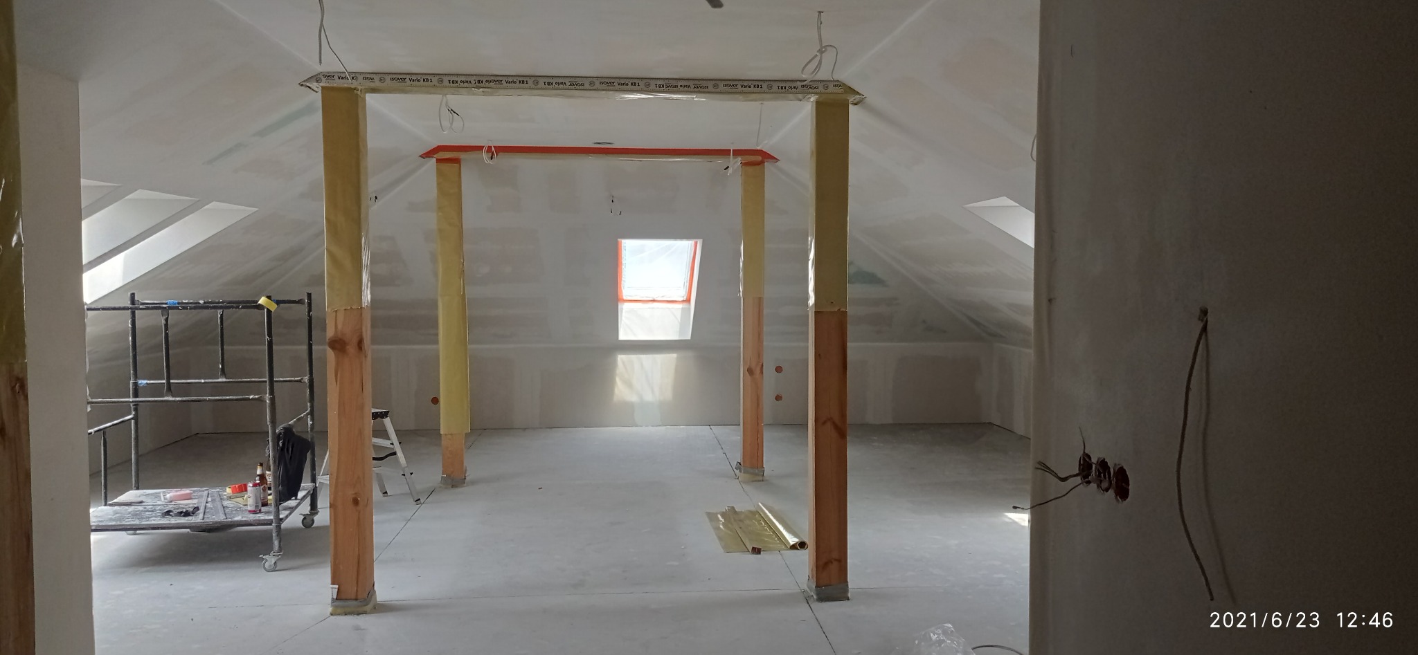 Poddasze w trakcie adaptacji: surowe drewniane słupy, częściowo zabezpieczone folią, podtrzymują konstrukcję. Widoczne okno dachowe i ściany przygotowane do dalszych prac.