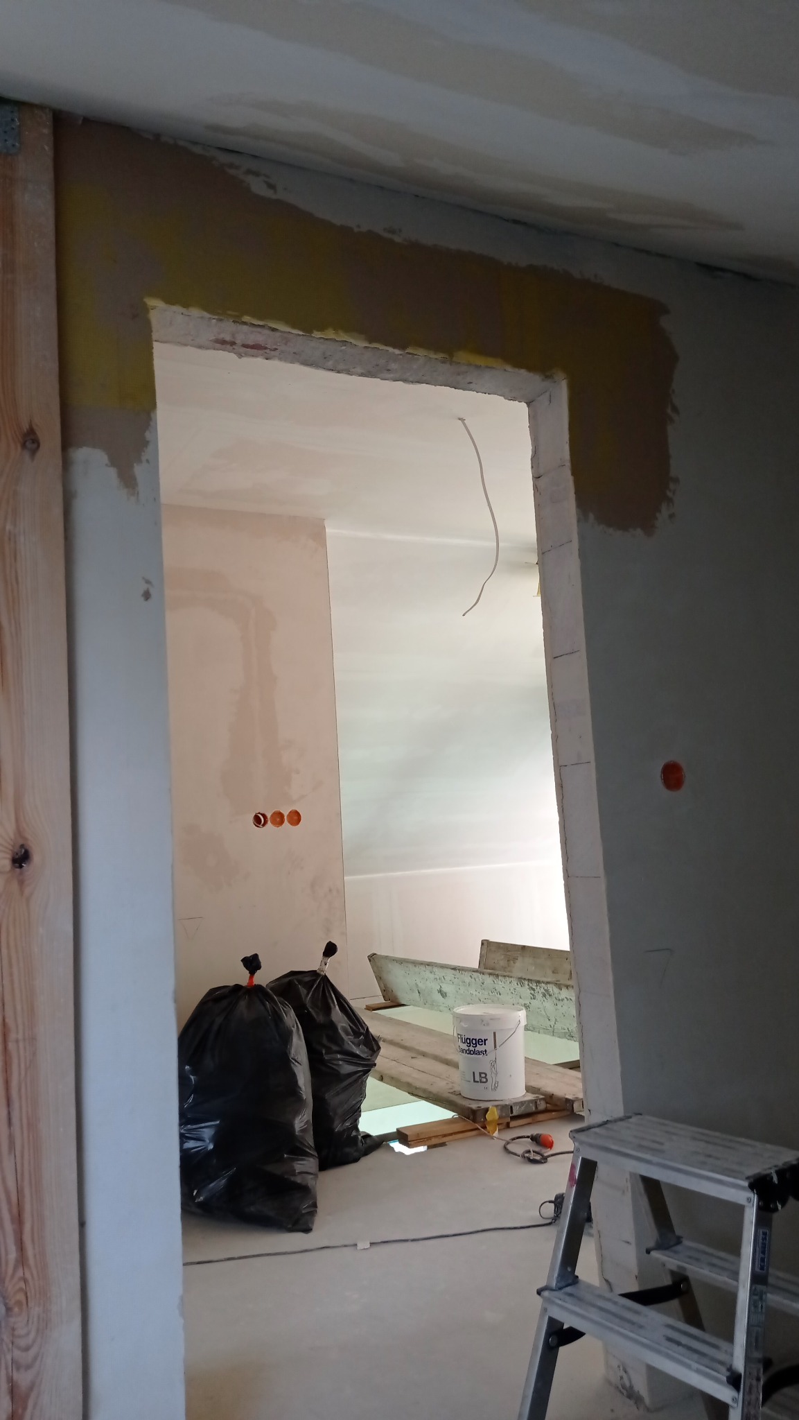 Wnętrze w trakcie prac wykończeniowych: widoczne szpachlowane ściany, przygotowane do malowania, z workami na gruz i narzędziami, w tym drabina i wiadro farby.