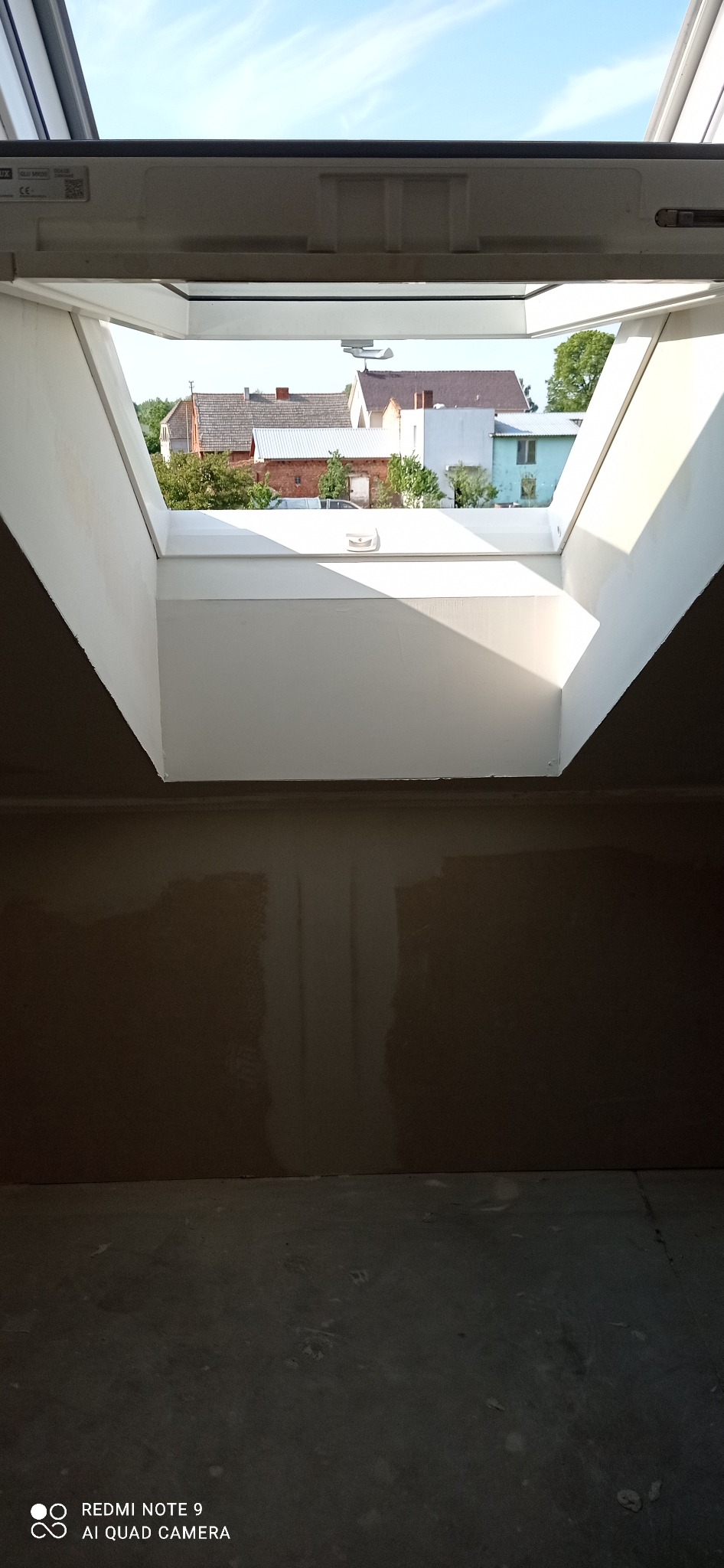 Wnętrze z oknem dachowym po szpachlowaniu, widok na zabudowę jednorodzinną. Ściany przygotowane do malowania, widoczne ślady po szpachli. Jasne niebo w tle.