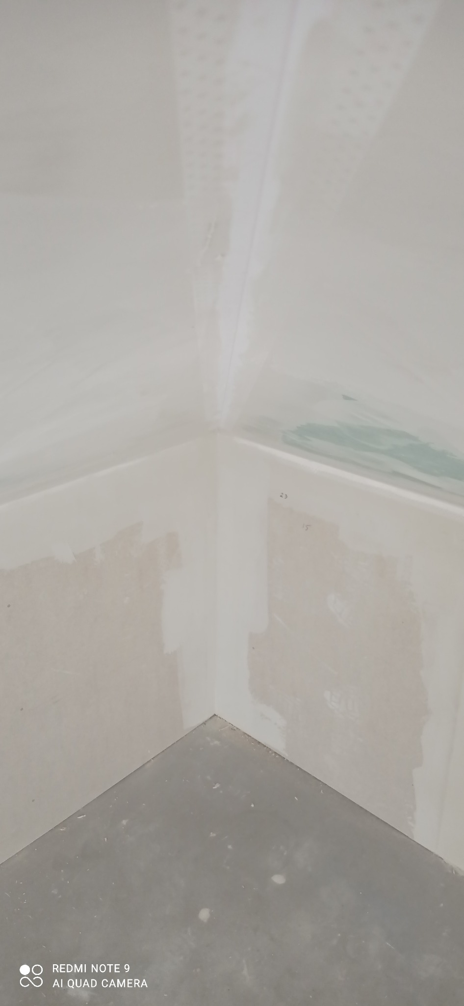 Narożnik pokoju w trakcie szpachlowania. Widoczne ślady szpachli na ścianach i suficie, surowy beton na podłodze. Przygotowanie do malowania.