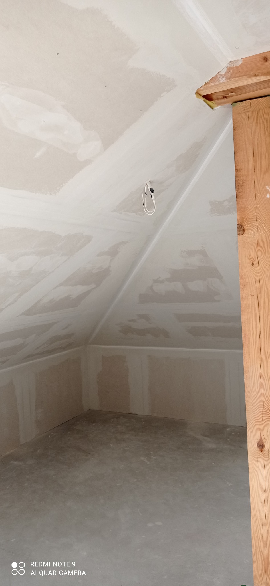 Poddasze po szpachlowaniu, widoczne łączenia płyt gipsowych na skośnym suficie i ścianach. Surowy drewniany słup w prawym rogu kadru.