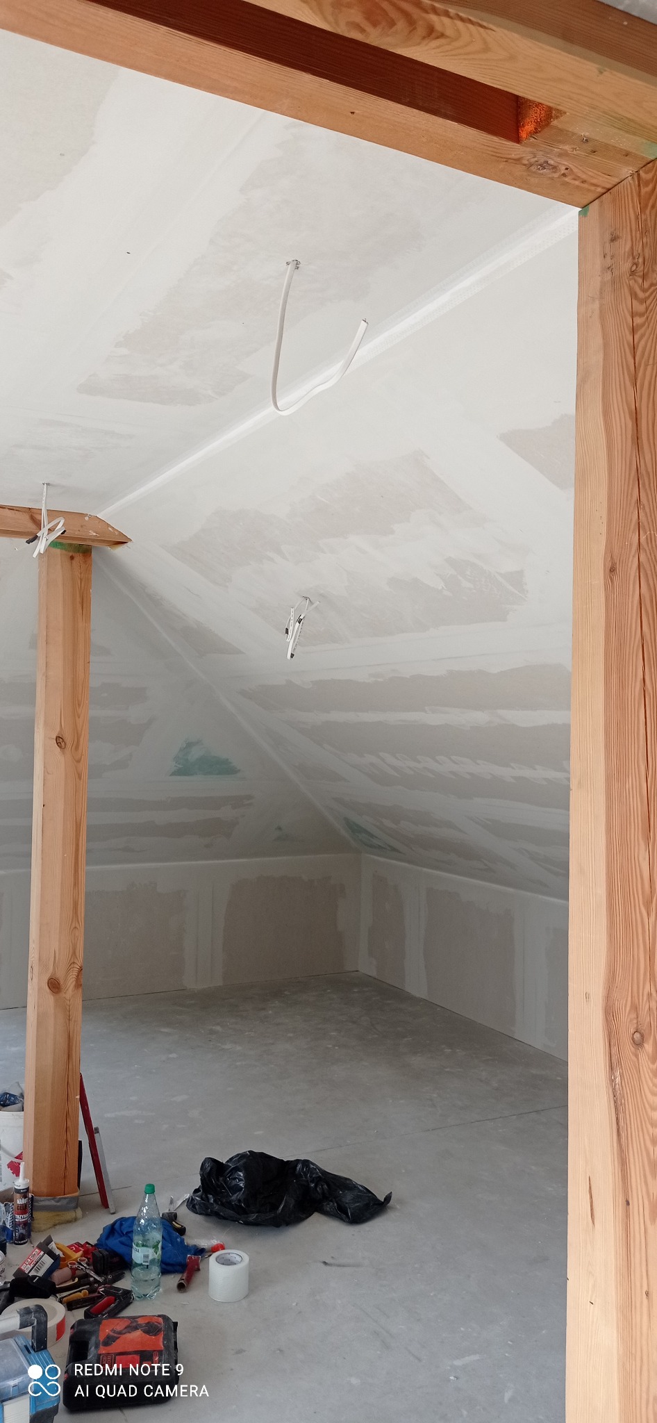 Poddasze w trakcie wykańczania: płyty karton-gips na skosach, drewniane elementy konstrukcyjne, widoczne przewody elektryczne, narzędzia i materiały budowlane na podłodze.