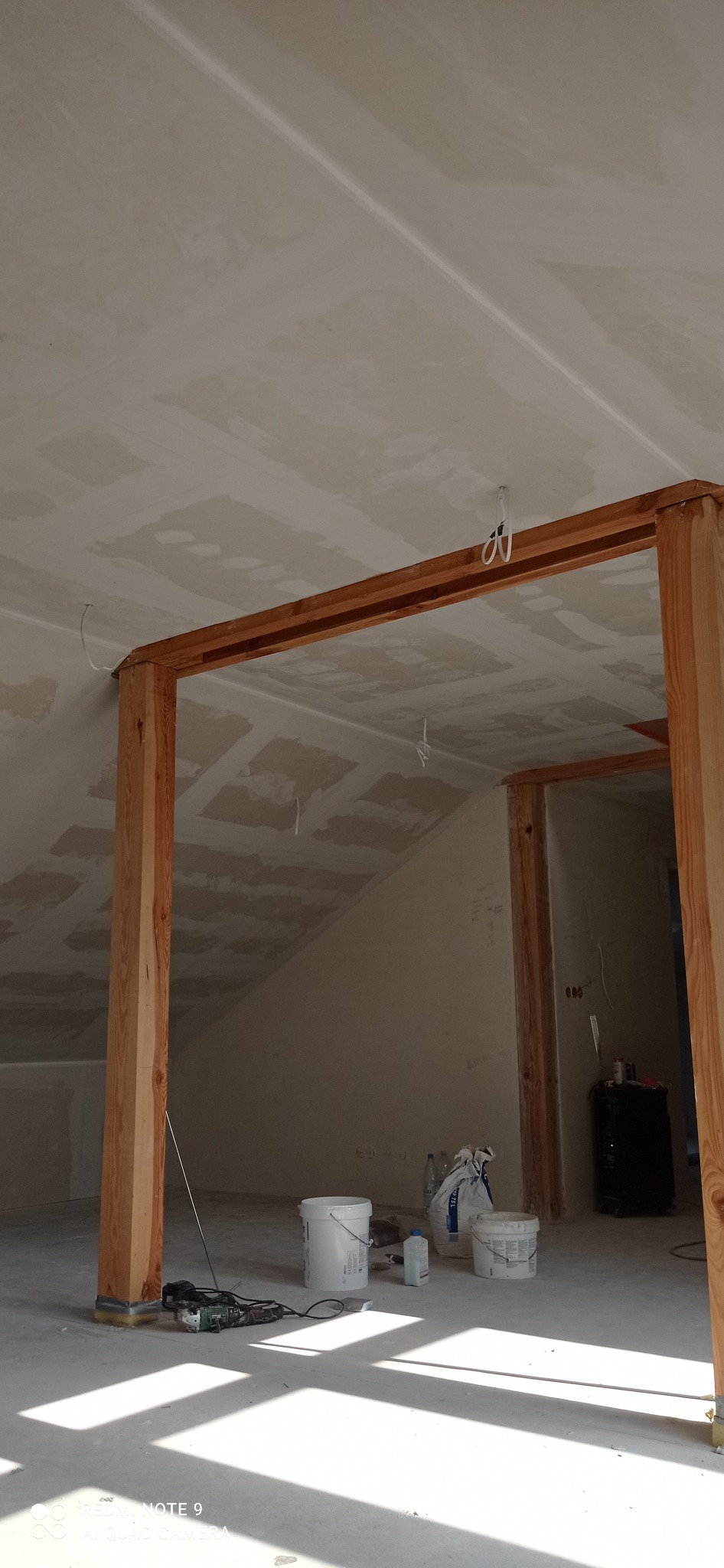 Poddasze w trakcie prac wykończeniowych: drewniana konstrukcja, szpachlowana sufit i ściany, narzędzia i materiały budowlane na podłodze. Widok z dołu, jasne światło.