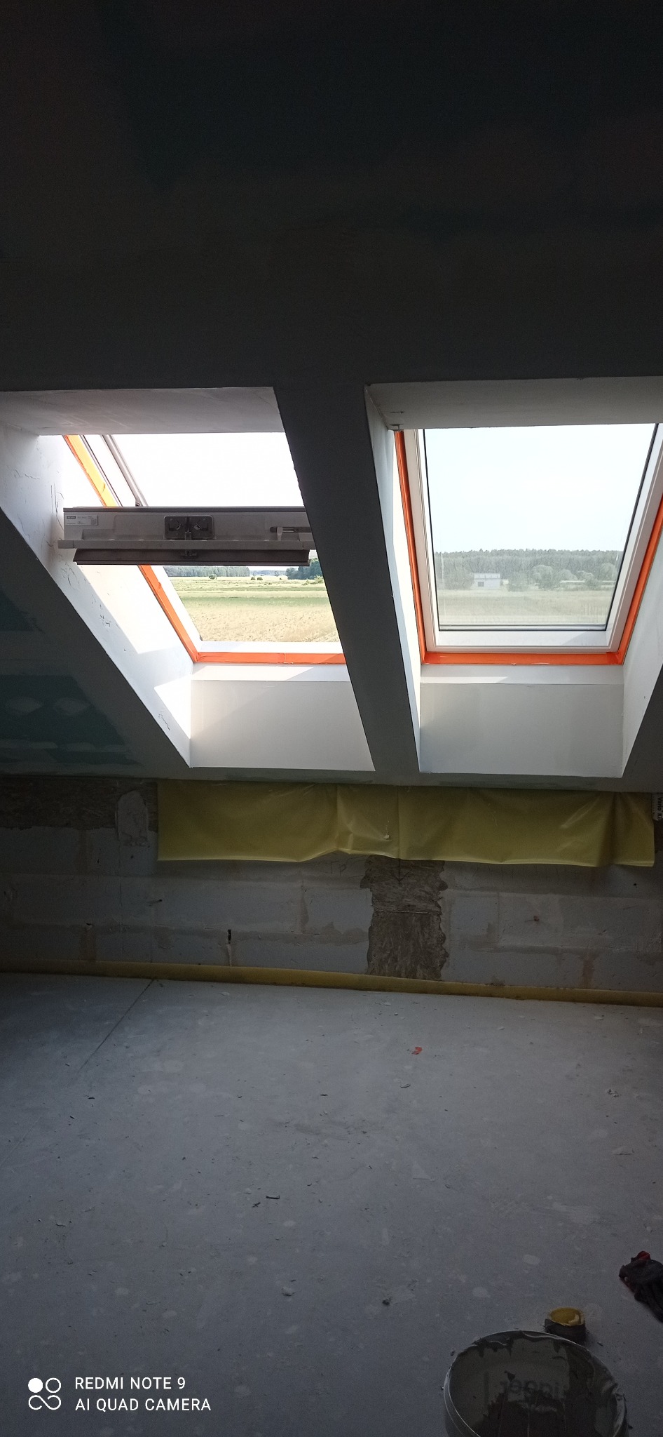 Dwa okna dachowe z pomarańczowymi ramami w surowym wnętrzu. Widok na pole przez okna. Ściany z bloków, częściowo zakryte folią. Podłoga betonowa, w tle wiadro.