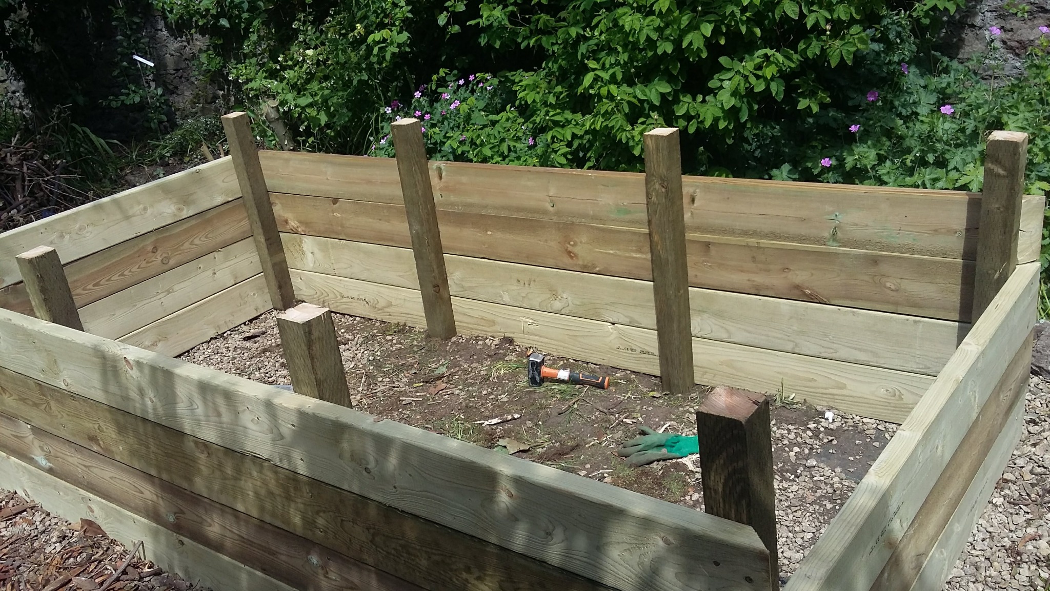 Drewniana konstrukcja skrzyni ogrodowej w trakcie montażu, widoczne deski, słupki, młotek i rękawice robocze. Tło stanowi zielona roślinność.