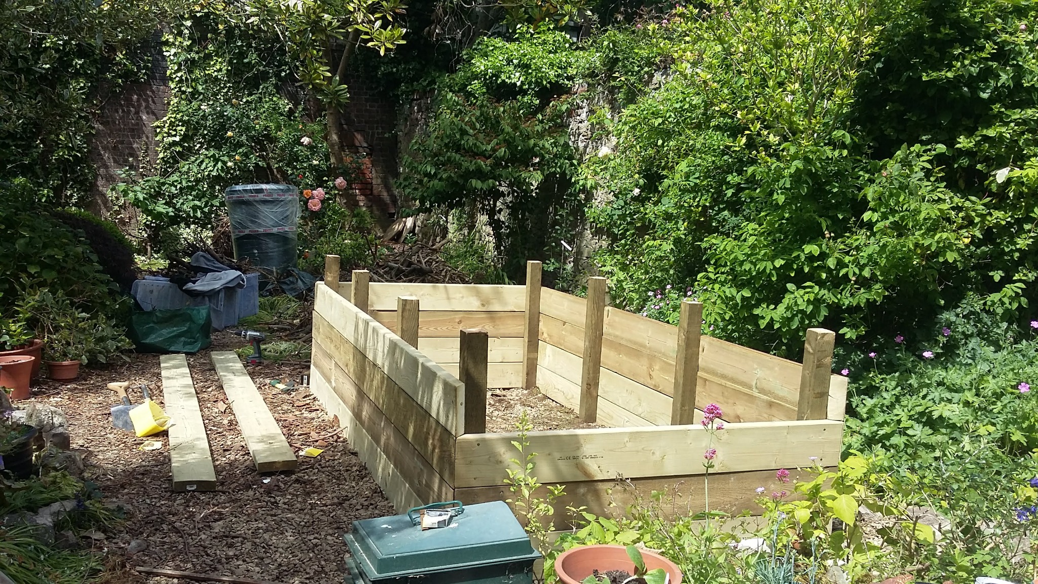 Drewniana konstrukcja podwyższonej grządki w ogrodzie, w trakcie montażu. Widoczne deski, słupki, narzędzia i bujna roślinność w tle. Ujęcie w słoneczny dzień.