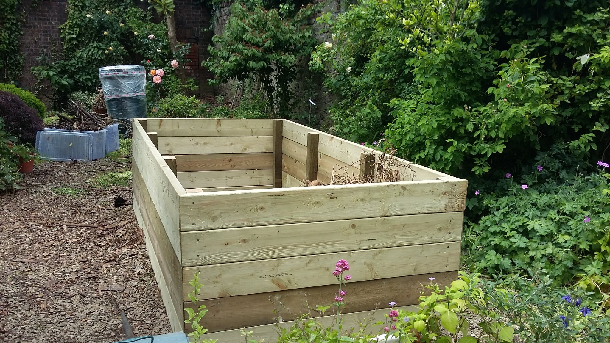 Drewniana konstrukcja skrzyni w ogrodzie, wykonana z jasnych desek, z widocznymi elementami mocującymi. W tle bujna roślinność i narzędzia ogrodnicze.