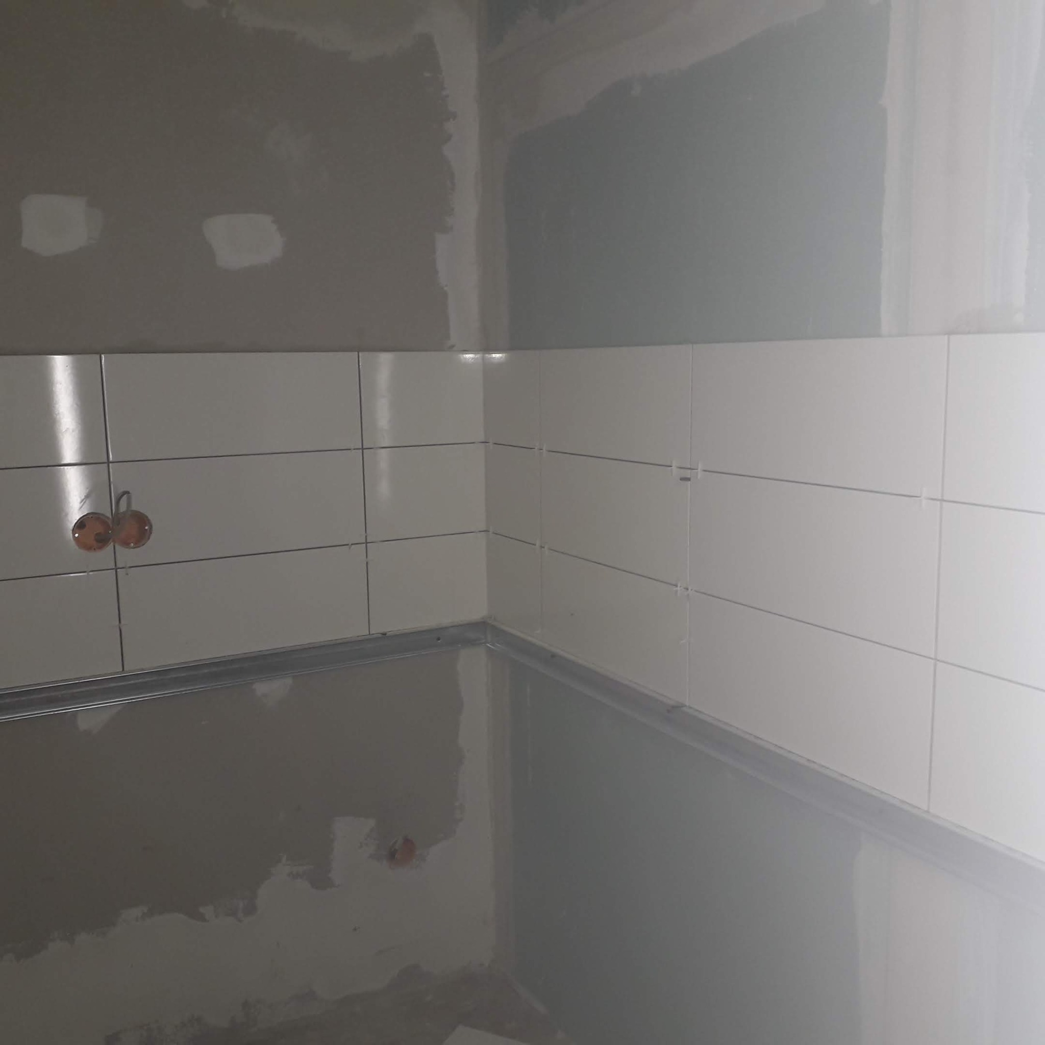 Fragment ściany z białymi płytkami, widoczne puszki elektryczne i niedokończone szpachlowanie powyżej. Ujęcie z bliska, ukazujące etap prac wykończeniowych.