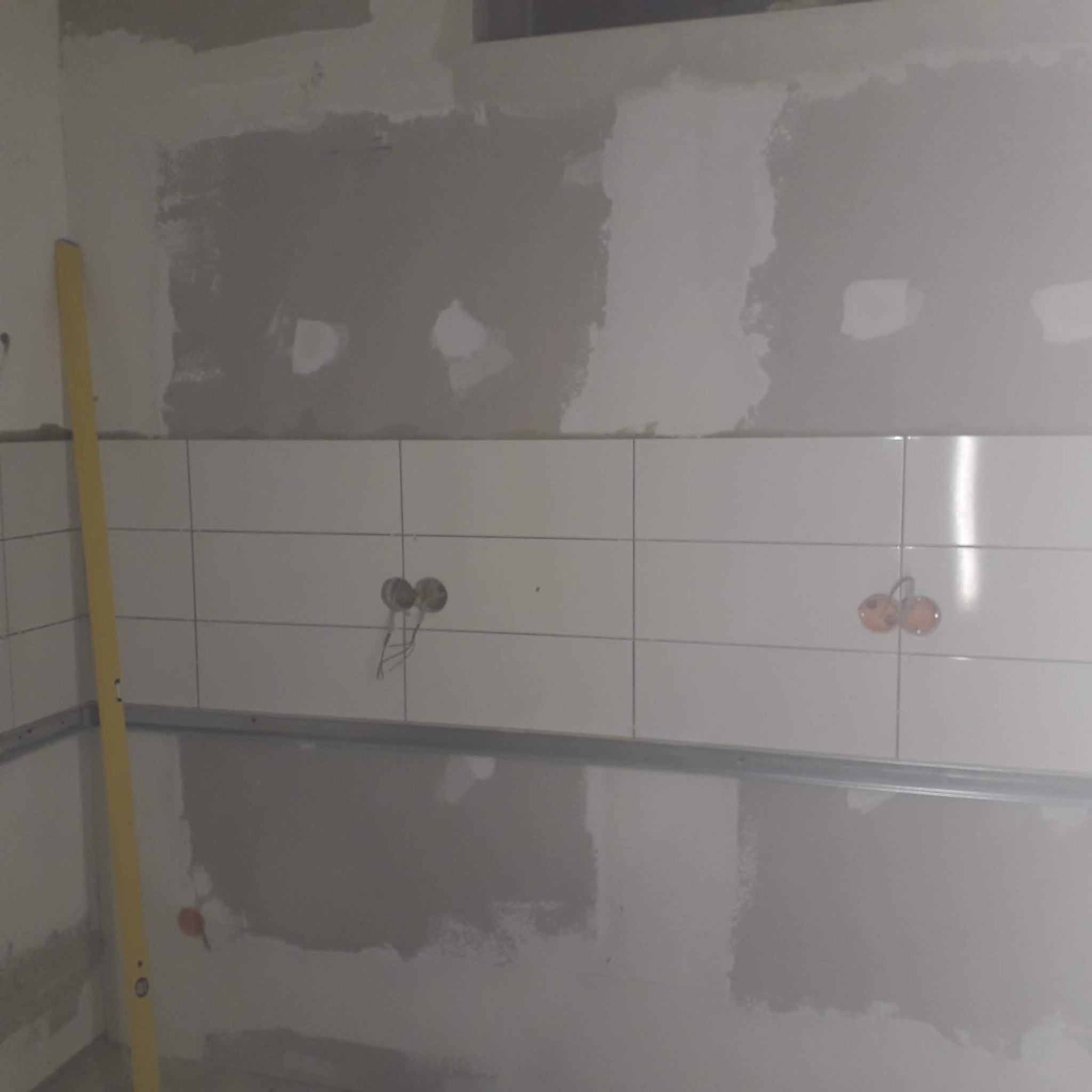 Ściana w trakcie wykańczania: białe płytki do połowy, powyżej szpachlowana powierzchnia. Widoczne puszki elektryczne i poziomica.