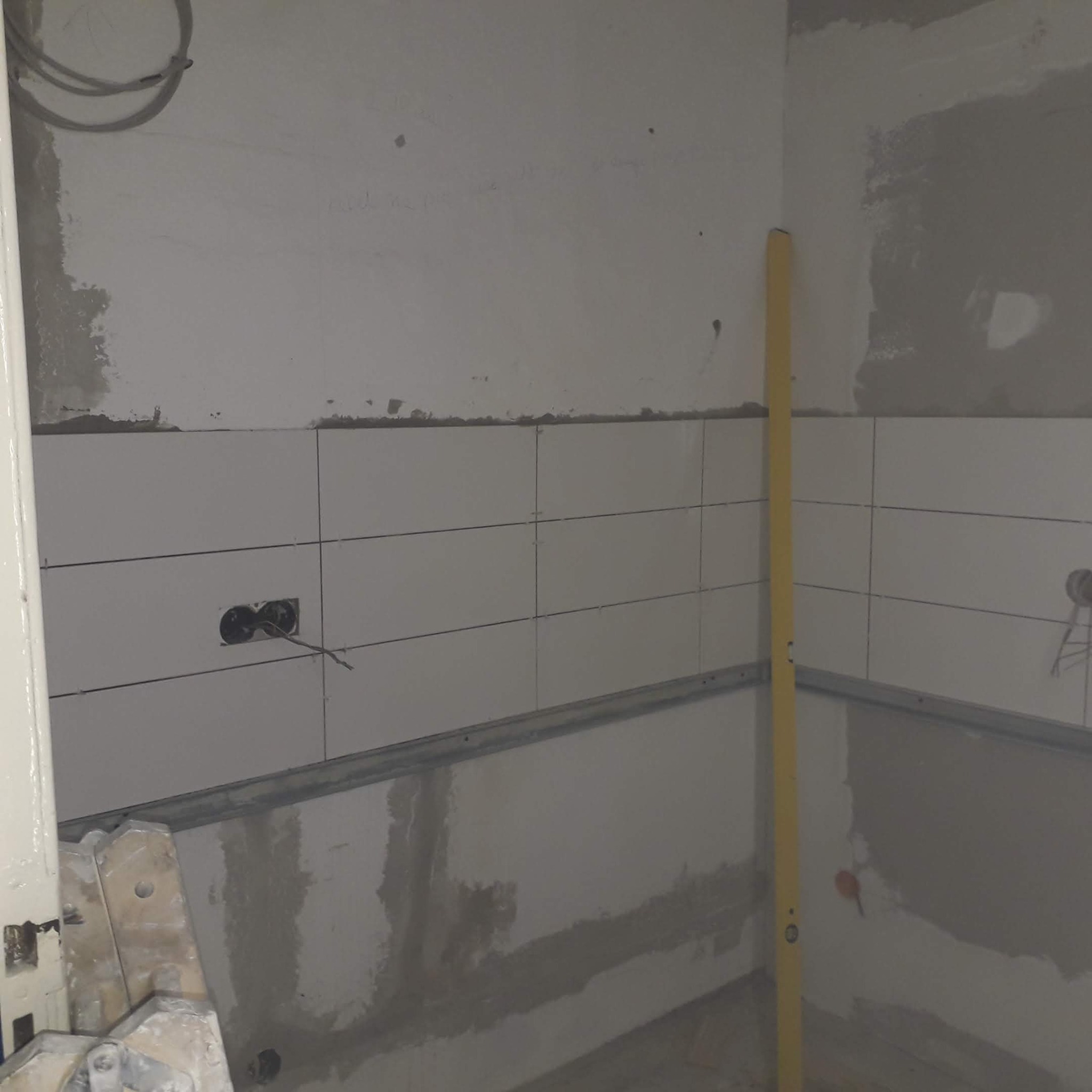 Ściana z częściowo położonymi białymi płytkami, widoczne gniazdka elektryczne i poziomica. Etap prac wykończeniowych w pomieszczeniu.