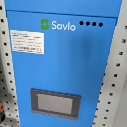 Wojciech Zaczek - Niebieski kompensator mocy biernej Savlo z panelem sterowania i tabliczką znamionową, zamontowany na ścianie z perforacją. Widoczne oznaczenia CE i dane techniczne.