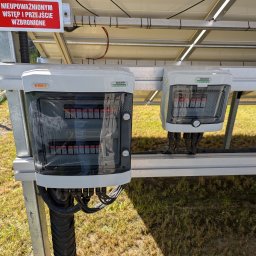 Wojciech Zaczek - Dwa białe rozdzielacze elektryczne KENO i NOARK zamontowane pod panelami słonecznymi, z widocznymi kablami i tabliczką ostrzegawczą.