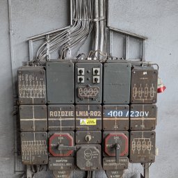 Wojciech Zaczek - Stara, przemysłowa rozdzielnia elektryczna z przewodami na szarej, betonowej ścianie. Widoczne oznaczenia, schematy i przełączniki. Charakterystyczny, techniczny detal.