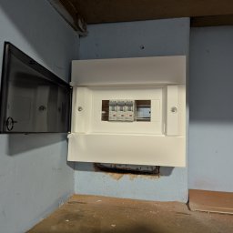 Wojciech Zaczek - Biała skrzynka rozdzielcza z bezpiecznikami Eaton, zamontowana na niebieskiej ścianie. Drzwiczki skrzynki otwarte po lewej stronie, widoczne trzy bezpieczniki.