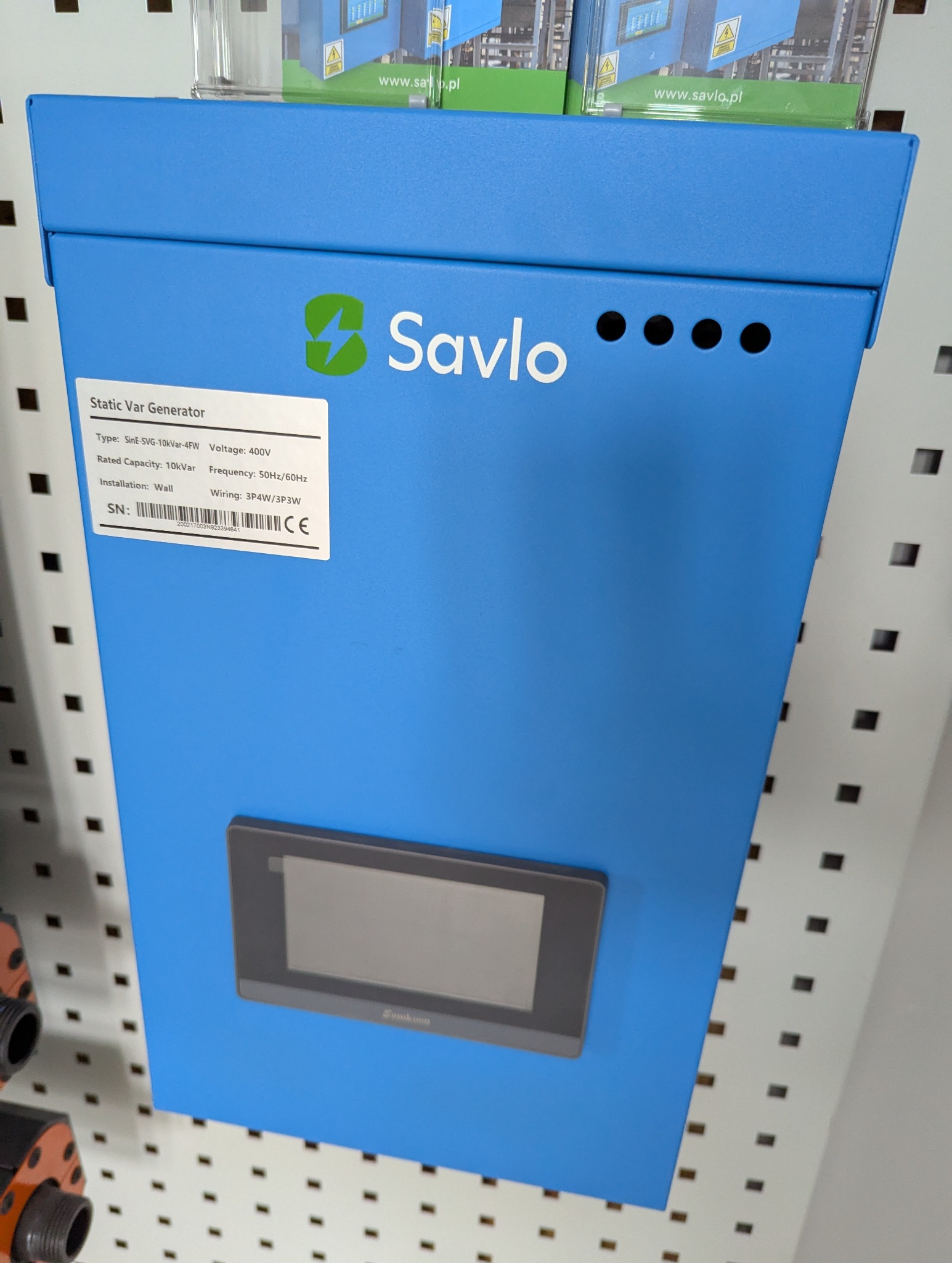 Niebieski kompensator mocy biernej Savlo z panelem sterowania i tabliczką znamionową, zamontowany na ścianie z perforacją. Widoczne oznaczenia CE i dane techniczne.